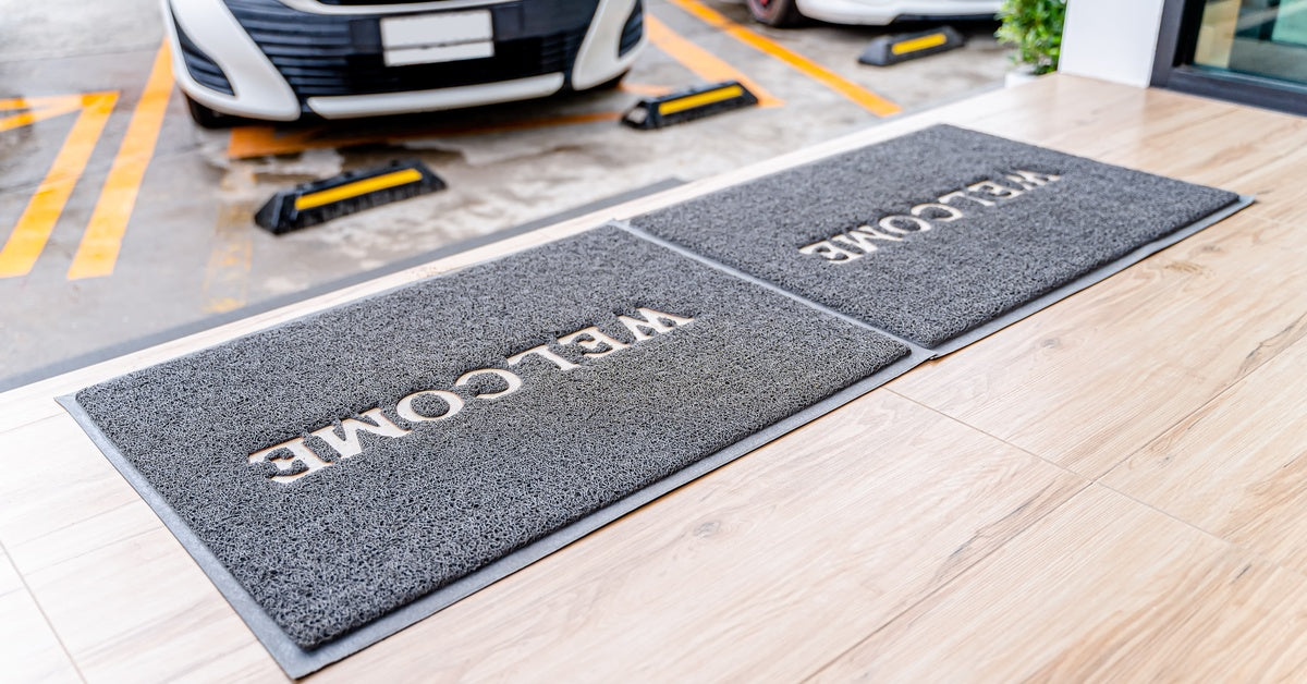 Two welcome mats are on the ground. IN the background, there are several cars in a parking lot.