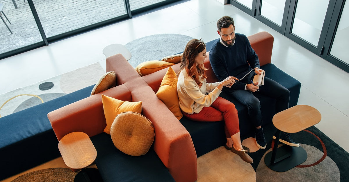 A man and woman sit on a couch next to one another. The couch has blue seat cushions and red back rests.