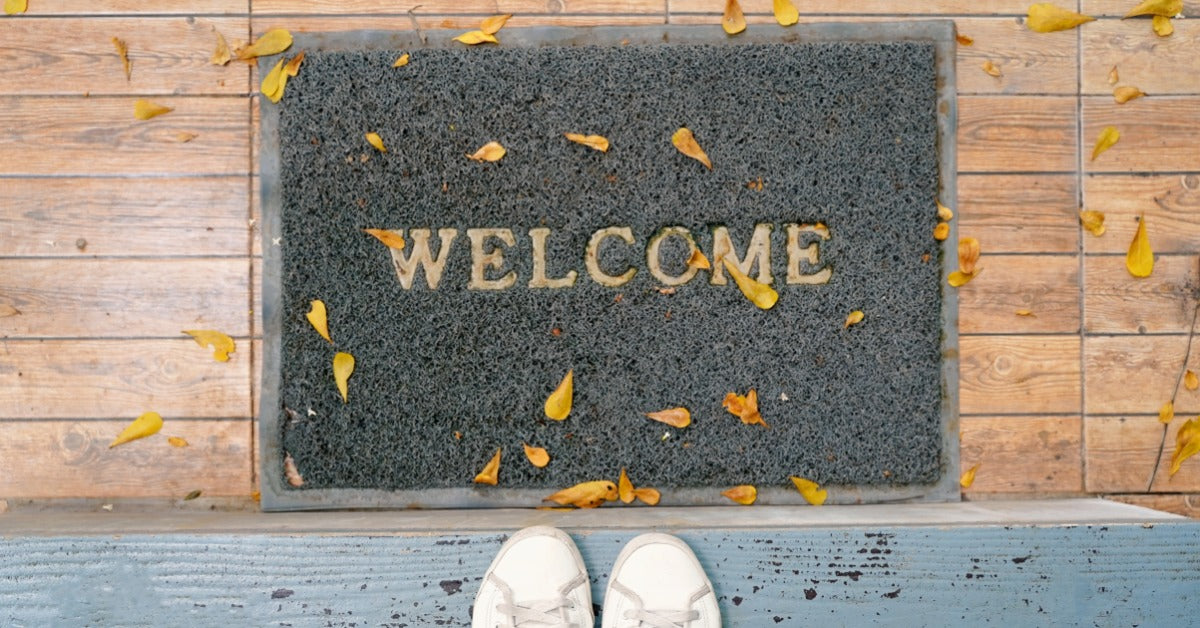 A person in white shoes is standing in front of a welcome mat. The mat is black with yellowish lettering.