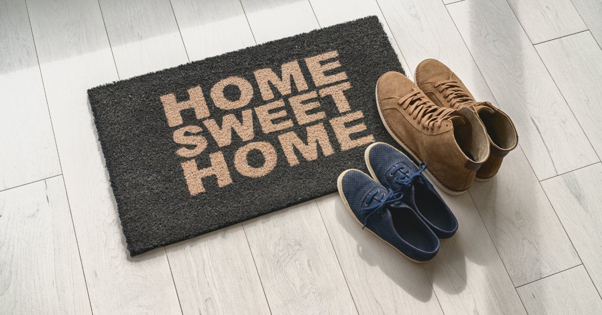 Two pairs of shoes, one pair is brown and the other is blue, are sitting on a welcome mat. The floor under the mat is gray tile.