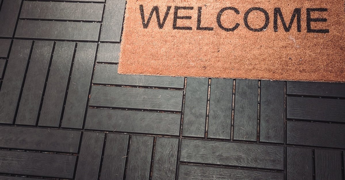 A welcome mat is placed on a deck that has dark wood. The mat is light brown in color and there is some dirt on it.