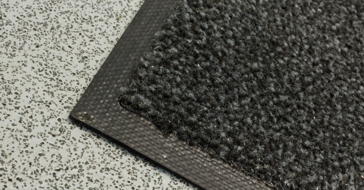 A black Berber mat is sitting on a floor. The floor underneath the mat is light gray with some darker texturing.