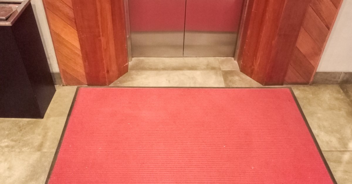 A red carpet is placed in front of an elevator door. There are wood columns on either side of the elevator.