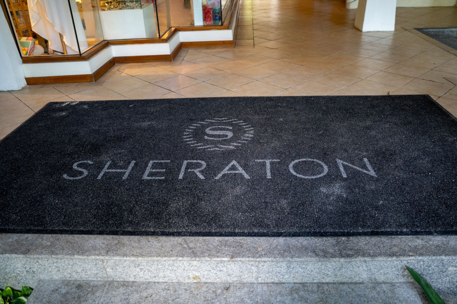 Closeup of a company’s black logo doormat at the entrance of a shop designed with their logo and the word Sheraton.