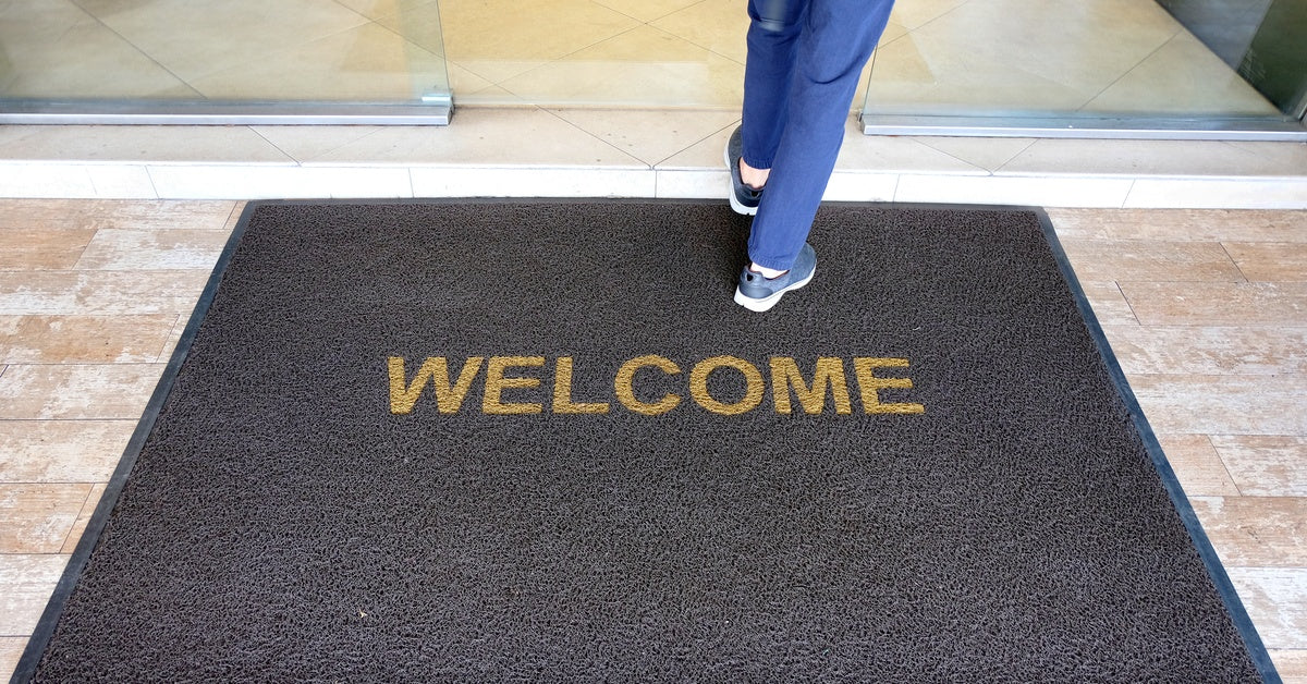 A welcome mat with the word "Welcome" on it at the entrance of a restaurant. A customer steps on the mat as she enters.