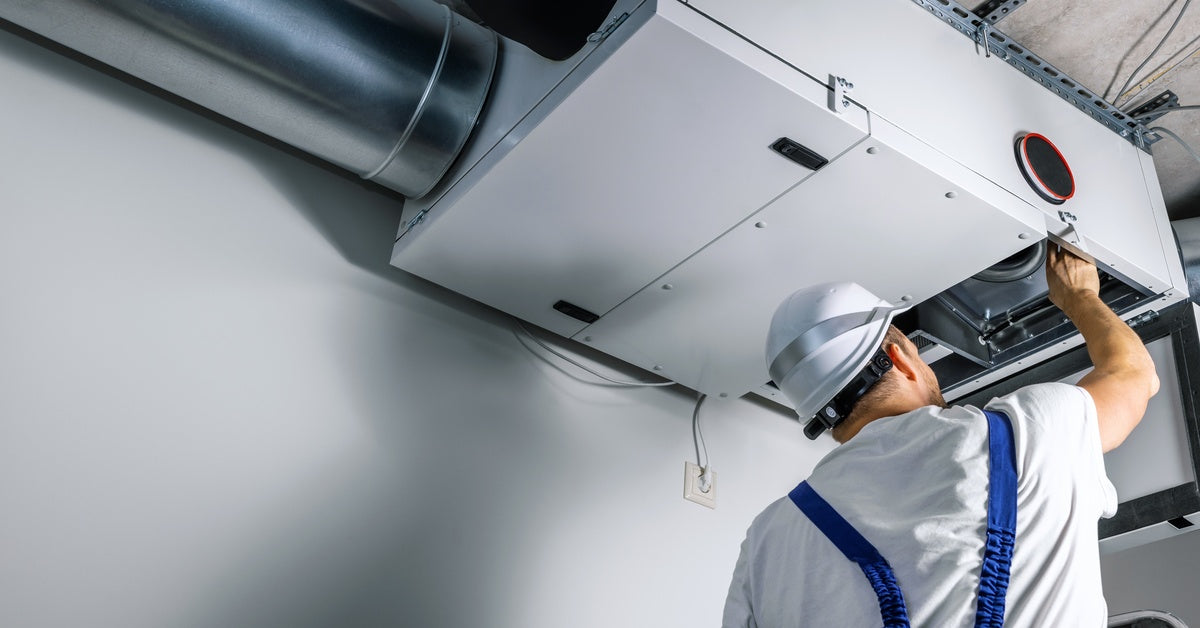 A man with blue overalls is sticking his hand in an HVAC system. The man is wearing a white hardhat.