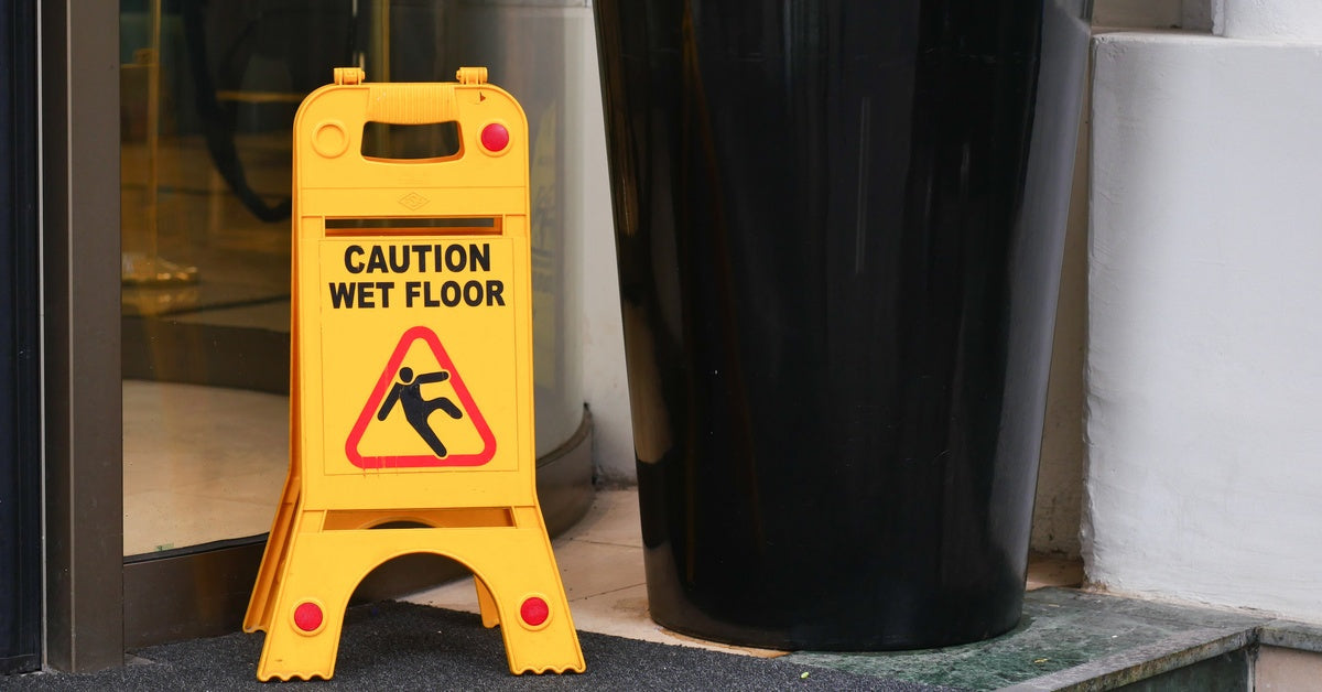 The entrance of a building where a dark mat sits in front of the door. There is a yellow "CAUTION WET FLOOR" sign on the mat.