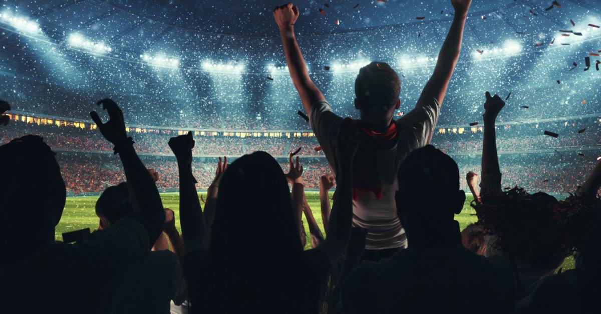 Confetti in the air and the silhouettes of several people with their arms up cheering in the stands at a stadium.