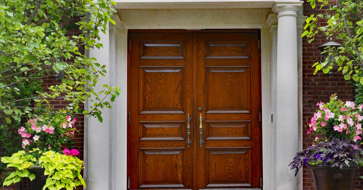 A set of wood double doors and a portico entrance to a brick house. Trees and potted plants are on each side.