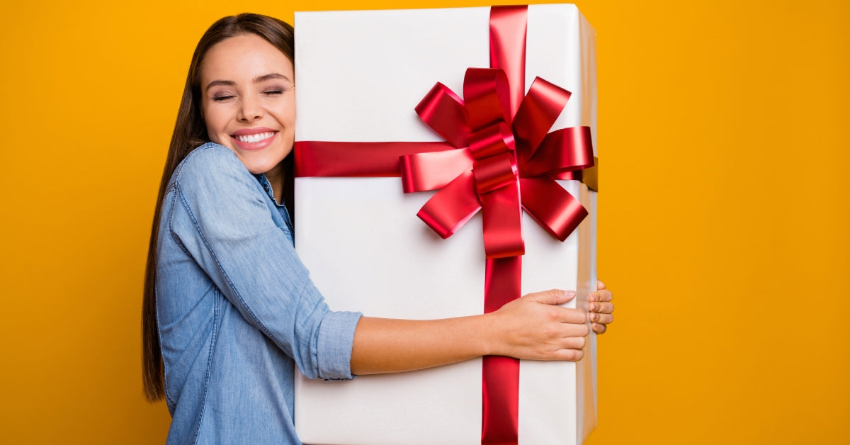 A brunette woman is hugging a large white box with a red ribbon in a bow in front of a yellow backdrop.