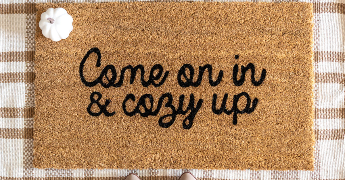 A base doormat with a brown check pattern sits beneath a coir doormat that says Come On In and Cozy Up in black cursive.