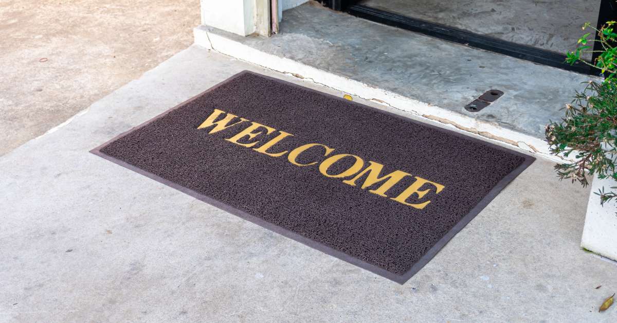 A rectangular doormat reading "welcome" in gold letters on a concrete entryway in front of a business doorway.