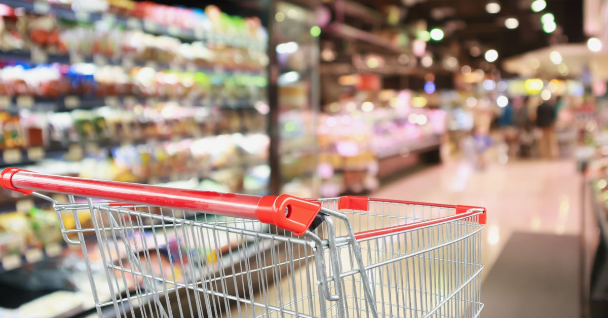 A shopping cart with a red plastic handle is in the foreground in a grocery store with blurred surroundings.