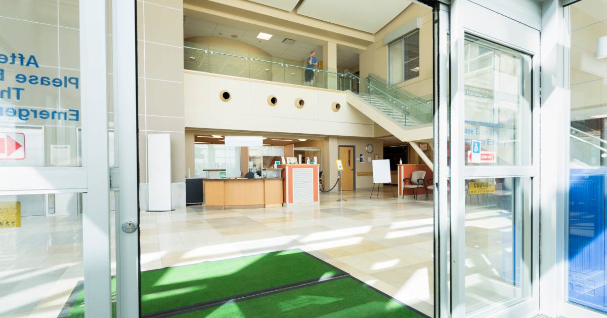 The entrance of a hospital lobby with sliding automatic glass doors and two carpeted green entrance mats.