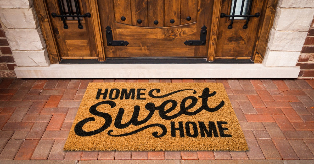 A coir doormat reading "home sweet home" on a brick porch in front of a wood door with side windows.