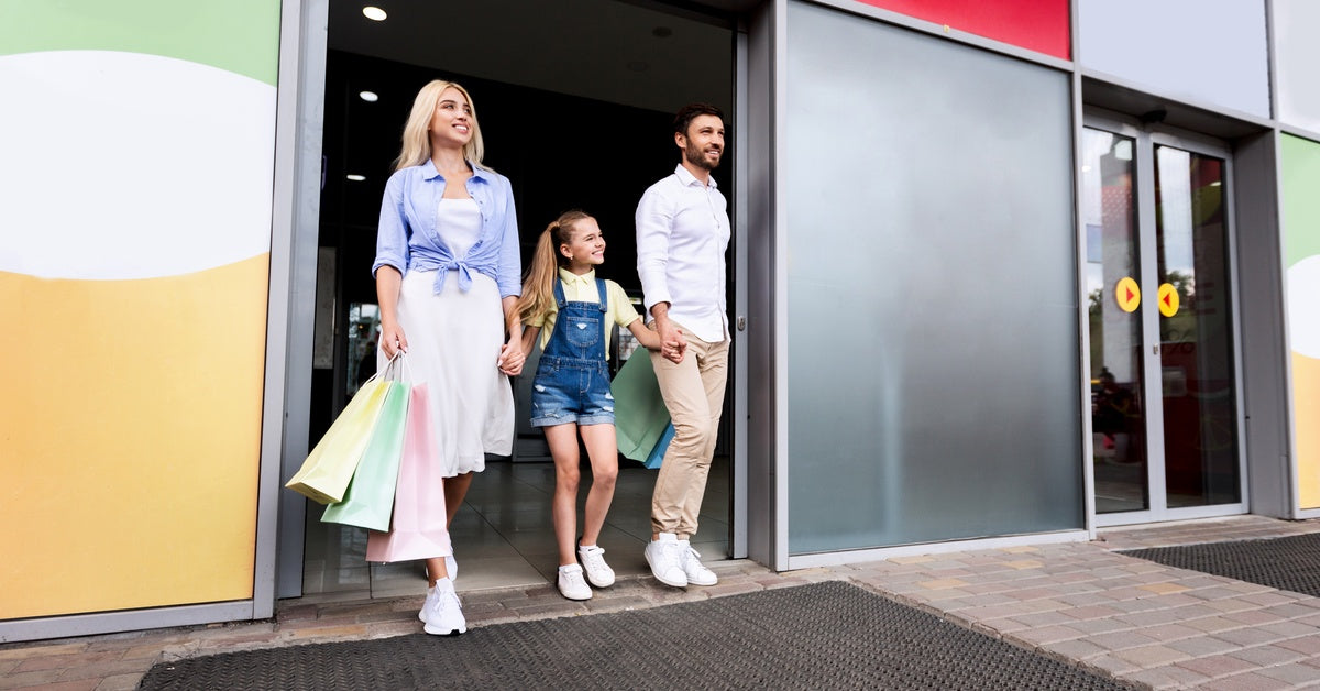 A couple holds hands with a young girl between them, carrying multicolored shopping bags while leaving a store.